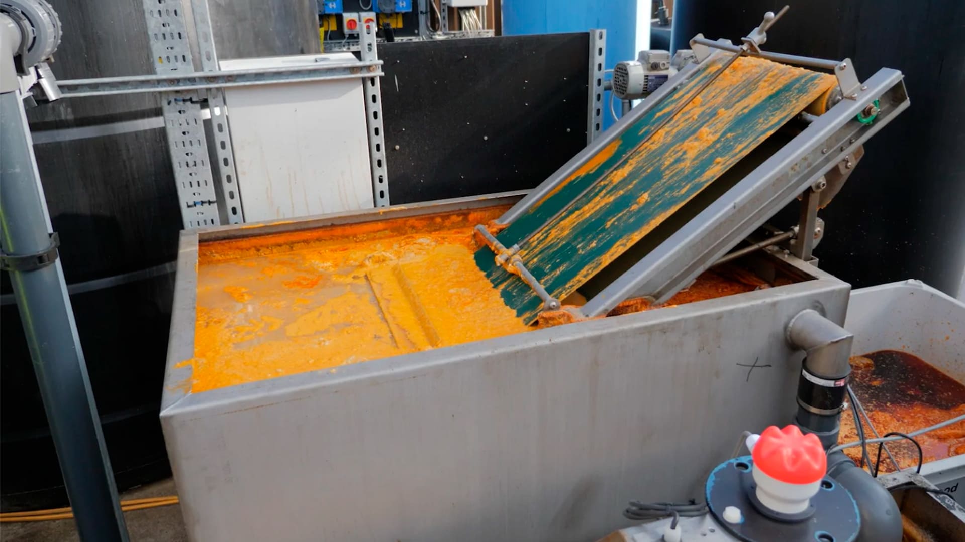 Camión rojo de plataforma larga transportando un gran decantador amarillo de la marca 'SYNERTECH' para el tratamiento de aguas residuales complejas, estacionado en una calle urbana con edificios y cielo despejado al fondo. - ver detalles