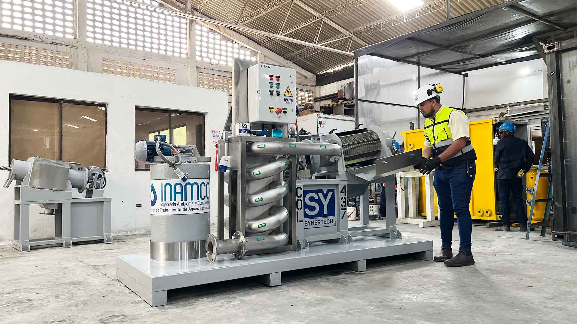 Técnico inspeccionando una planta de tratamiento de aguas residuales modular de color verde marca BIOMAX SYNERTECH, que incluye módulos de pretratamiento, decantación, DAF y oxidación avanzada. La planta está ubicada en un entorno industrial con techo alto y tuberías visibles. - ver más información
